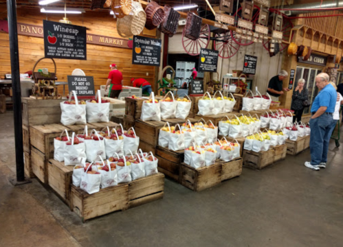 A market display with wooden crates filled with bags of apples, surrounded by shoppers and vendors in a rustic setting.