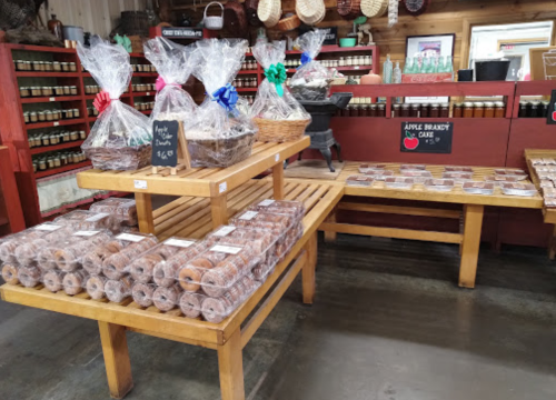 A rustic shop display featuring baskets of treats and jars, with a table of packaged goods and a sign for apple brandy cake.