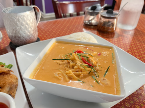 A bowl of creamy curry with bamboo shoots and red peppers, served with a side of rice on a patterned tablecloth.