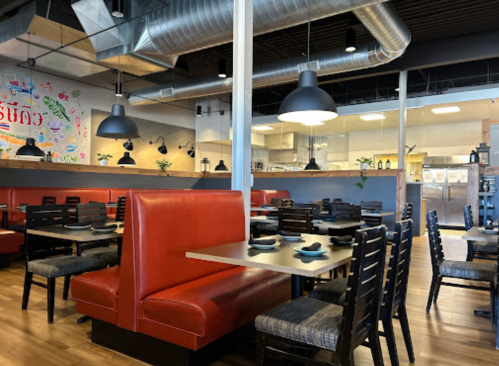 Interior of a modern restaurant featuring red booths, wooden tables, and a visible kitchen area in the background.