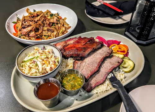 A plate with sliced brisket, coleslaw, pickles, and sauces, alongside a bowl of stir-fried noodles with vegetables.