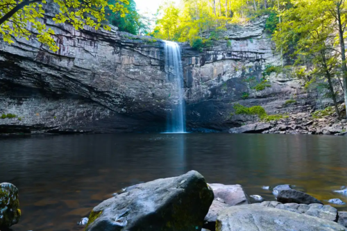 A serene waterfall cascades into a calm pool, surrounded by rocky terrain and lush green trees.