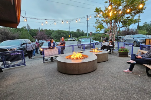 Outdoor seating area with fire pits, string lights, and people socializing in a lively atmosphere.