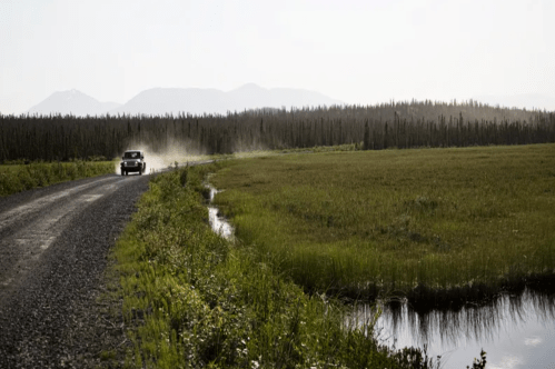 A vehicle drives along a gravel road beside a grassy field and a small pond, with mountains in the background.