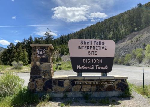 Sign for Shell Falls Interpretive Site in Bighorn National Forest, featuring stone base and forested background.