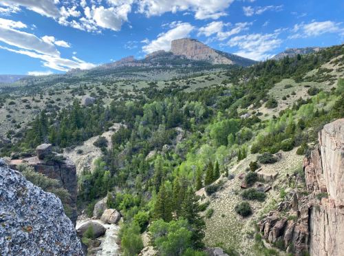 A scenic view of a mountainous landscape with green trees, rocky cliffs, and a blue sky with scattered clouds.