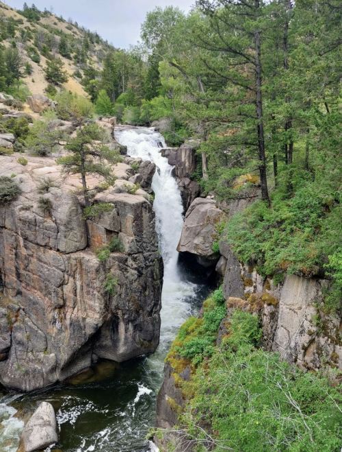 A scenic waterfall cascades between rocky cliffs, surrounded by lush greenery and trees in a natural landscape.