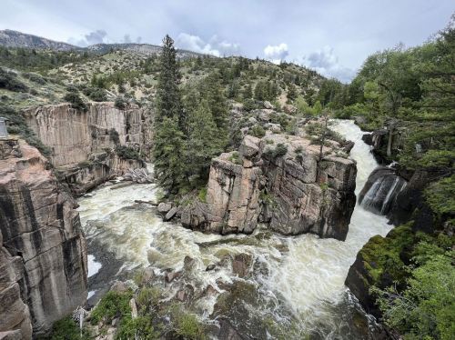 A rushing river flows between rocky cliffs, surrounded by lush greenery and mountains under a cloudy sky.
