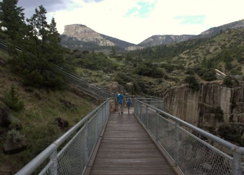 Two people walk on a wooden bridge surrounded by lush greenery and rocky cliffs under a cloudy sky.