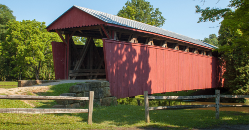 A red covered bridge with a metal roof, surrounded by trees and grass, spans a small stream.