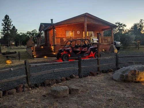 A log cabin with a porch, surrounded by trees, and an off-road vehicle parked in front on a gravel driveway at dusk.