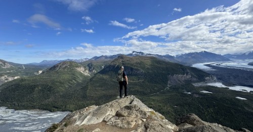 Hiker on a mountain summit overlooks the vast Matanuska Valley and a distant glacier on a sunny day.