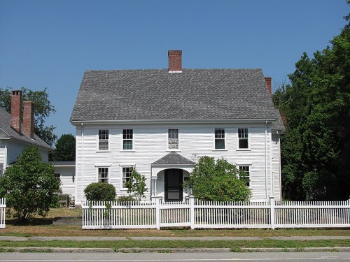 A white two-story house with a gray roof, surrounded by greenery and a white picket fence on a clear day.