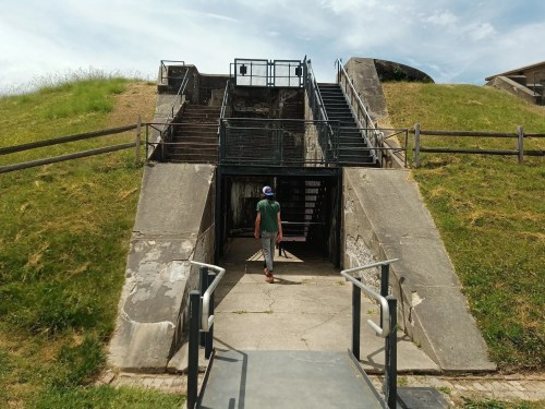 A person walks towards a concrete entrance with stairs leading up, surrounded by grassy hills and a wooden fence.
