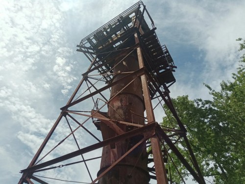 Rusty industrial tower against a cloudy sky, surrounded by greenery.
