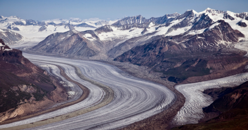 Aerial view of a winding glacier surrounded by rugged mountains and snow-capped peaks under a clear blue sky.