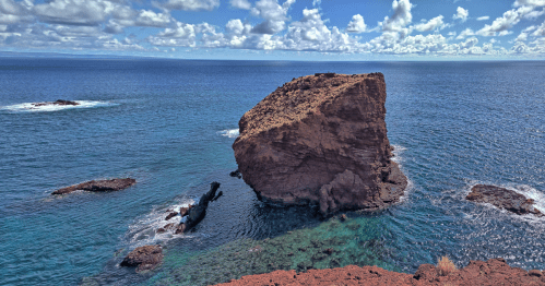 A large rock formation rises from the clear blue ocean, surrounded by smaller rocks and a vibrant sky with clouds.