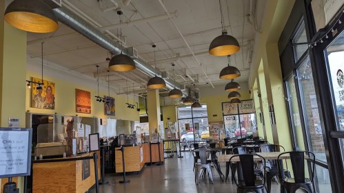 Interior of a bright café with yellow walls, metal chairs, and pendant lights, featuring artwork and a counter area.