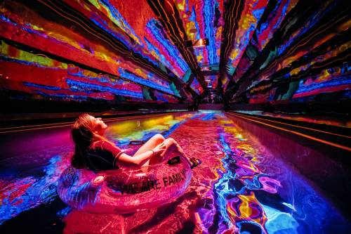 A person relaxes in a colorful pool on a float, surrounded by vibrant, swirling lights reflecting on the water.