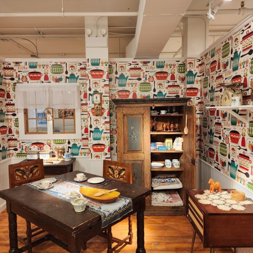 A cozy vintage kitchen with patterned wallpaper, a wooden table, and an antique cabinet filled with dishes and decor.