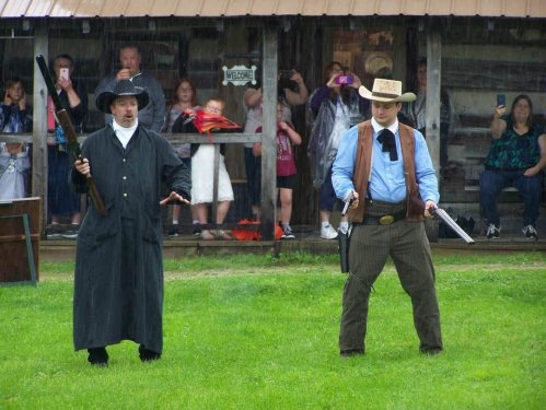Two men in Western attire face off in the rain, while a crowd watches from a covered area behind them.
