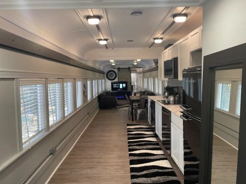 Interior of a converted bus featuring a modern kitchen, dining area, and workspace with large windows and stylish decor.