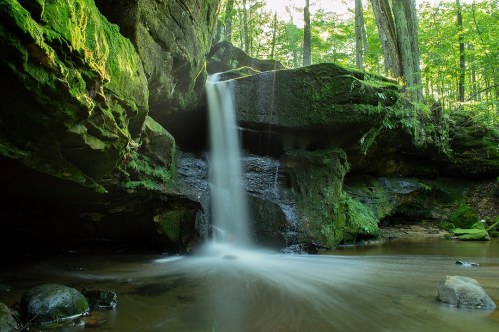 A serene waterfall cascades over moss-covered rocks in a lush green forest, creating a tranquil scene.