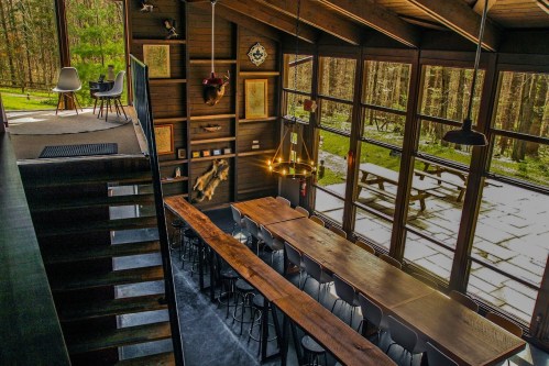 A spacious, modern dining area with large windows overlooking a forest, featuring a long wooden table and stylish chairs.