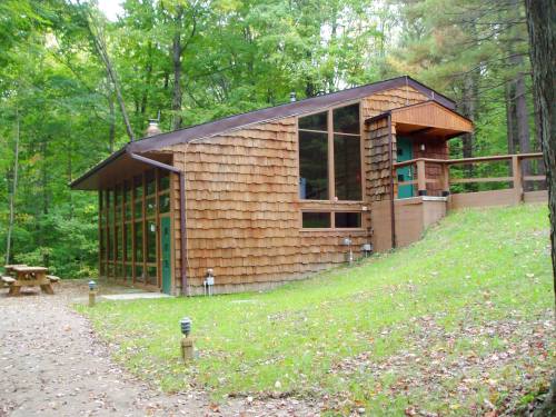 A modern cabin with wooden shingles, large windows, and a deck, surrounded by lush green trees and a gravel path.