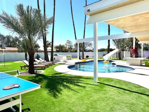 A sunny backyard with a pool, palm trees, lounge chairs, and a slide, featuring a ping pong table on green grass.