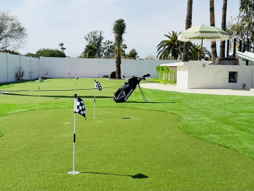 A putting green with small flags, a golf bag, and a shaded seating area surrounded by palm trees.