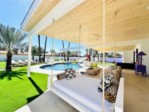 A stylish outdoor lounge area with a swing, pool, and palm trees, set in a sunny, modern backyard.
