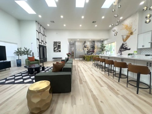 Spacious modern living area with a green sofa, bar stools, and artistic wall decor, featuring large windows and wooden flooring.