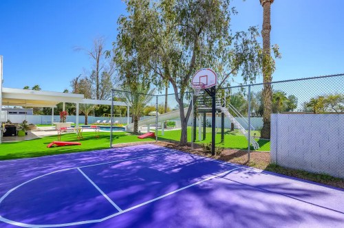 A vibrant outdoor basketball court with purple flooring, surrounded by greenery and a sunny blue sky.