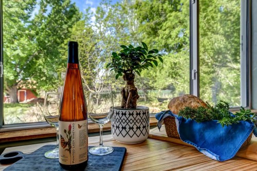 A bottle of wine, two glasses, a potted plant, and a loaf of bread on a wooden table by a sunny window.