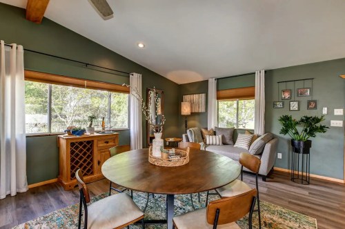Cozy living space with a round dining table, wine rack, and a comfortable sofa by large windows with greenery outside.