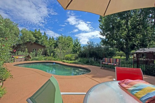 A sunny backyard with a pool, surrounded by greenery and a patio table with colorful towels.