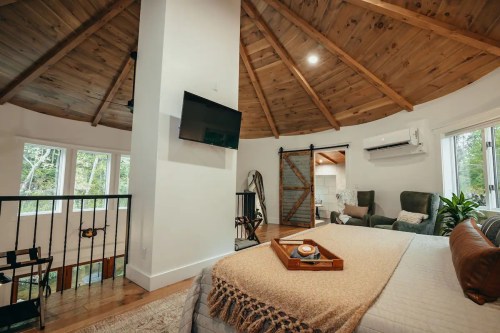 Cozy bedroom with a circular design, wooden ceiling, TV, and a view of greenery through large windows.