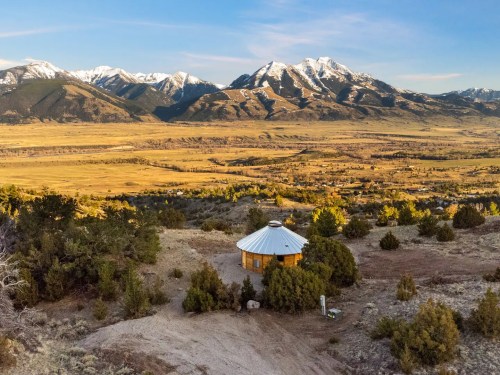 A scenic view of a yurt surrounded by trees, with snow-capped mountains and a valley in the background.
