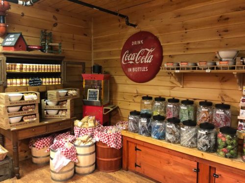 A rustic store interior with wooden walls, shelves of jars, and vintage Coca-Cola signage. Baskets and barrels are displayed.
