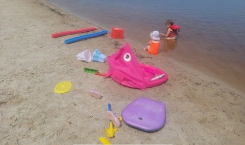 A sandy beach with colorful toys scattered around, and two children playing near the water's edge.