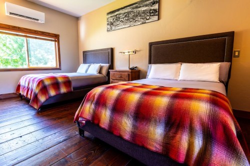 Cozy hotel room with two beds, wooden floors, and a large window letting in natural light. Colorful plaid blankets.