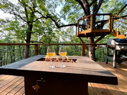Two glasses of wine sit on a fire pit table, surrounded by trees and a deck with a grill in a serene outdoor setting.