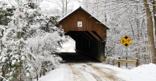 A snowy road crosses a wooden covered bridge surrounded by snow covered trees.