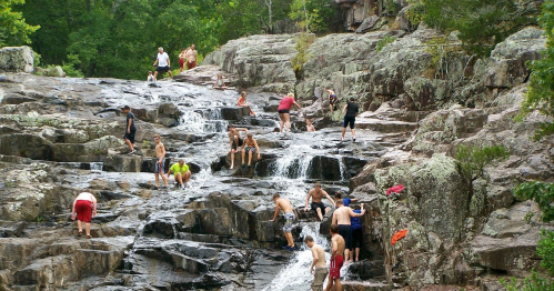 A group of people climbing and playing on rocky terrain near a waterfall in a lush, green forest.