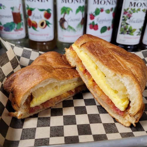 A croissant sandwich filled with egg and bacon, cut in half, served on a checkered paper with flavored syrups in the background.