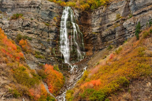A stunning waterfall cascades down rocky cliffs, surrounded by vibrant autumn foliage in warm hues.