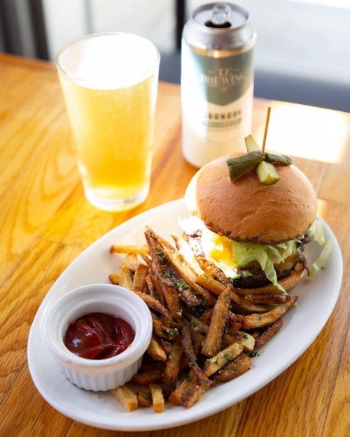 A plate with a burger topped with pickles, served with fries and ketchup, alongside a glass of beer.