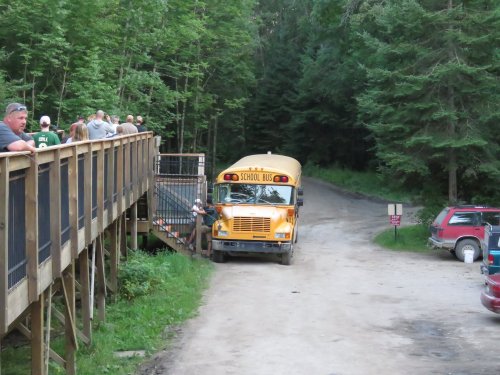 A yellow school bus parked on a dirt road near a wooden deck, surrounded by trees and people in the background.