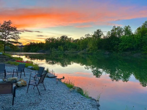 A serene lakeside scene at sunset, with colorful skies reflecting on calm water and chairs arranged on the shore.
